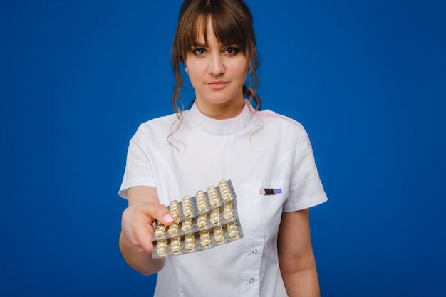 Het concept van gezondheidszorg. Een jonge brunette arts in een witte jas op een blauwe achtergrond toont platen met capsules om in te nemen.