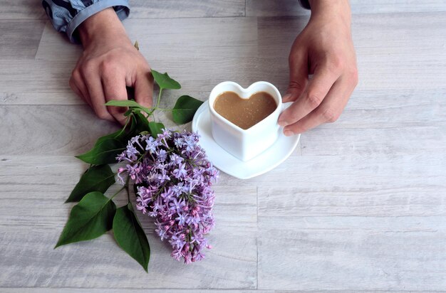 Het concept van goedemorgen Een kopje koffie in de vorm van een hart een tak van lila in mannenhanden op een grijze achtergrond bovenaanzicht