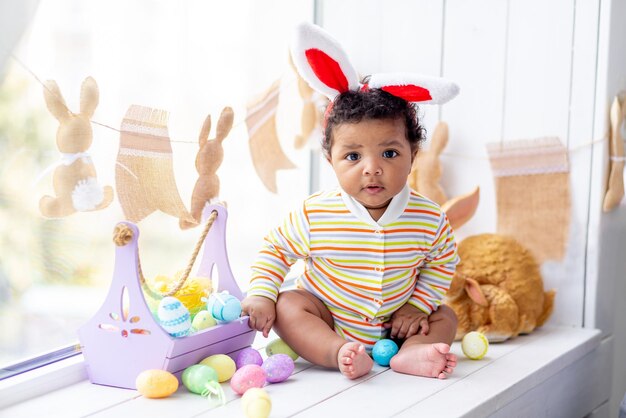 Het concept van Pasen een Afrikaans-Amerikaans klein kind in de oren van een haas op het hoofd met eieren en een konijn op het raam van het huis