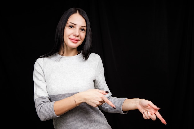 Het concept van reclame. Positieve, mooie, blanke vrouw met een stralende glimlach en lichte trui op een zwarte ruimte wijst op twee wijsvingers en kijkt naar de lege kopie ruimte.