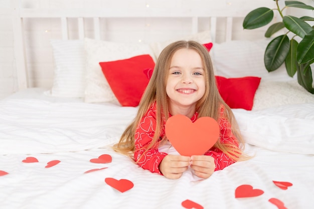 Het concept van Valentijnsdag een lief kindmeisje op een wit bed thuis in rode pyjama's tussen harten met een valentijnshart in haar handen glimlachend met geluk feliciteren met de vakantie