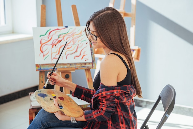 Het creatieve peinzende schildersmeisje schildert een kleurrijk beeld op canvas met olieverf in workshop.