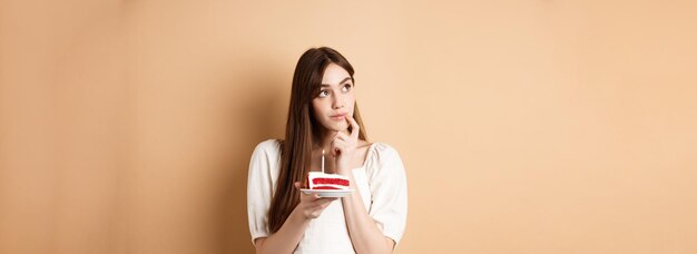 Het dromerige feestvarken kijkt opzij en denkt aan wens blazende kaars op cake die zich op beige backgr bevindt