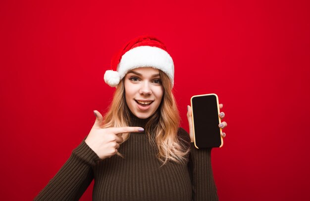 Het emotionele meisje in santahoed toont vinger op smartphone met het scherm