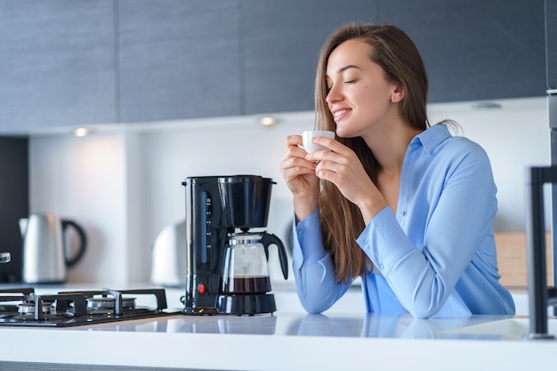 Het gelukkige aantrekkelijke vrouwelijke genieten van van vers koffiearoma na het brouwen van koffie thuis gebruikend koffiezetapparaat in de keuken. Koffieblender en huishoudelijke keukenapparatuur voor het maken van warme dranken