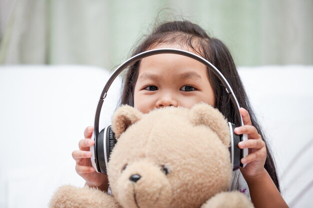 Het gelukkige Aziatische kindmeisje spelen met haar teddybeer met hoofdtelefoons in haar ruimte