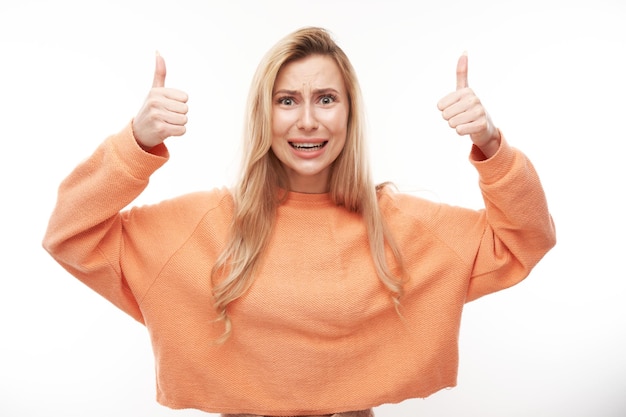 Het gelukkige blondemeisje toont hand met omhoog duimen en glimlacht bij de camera die op witte achtergrond wordt geïsoleerd