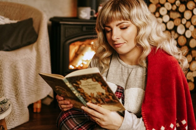 Het gelukkige boek van de vrouwenlezing voor Kerstmisbinnenland met open haard. Het meisje is gekleed in huiselijke, knusse kleren met een rode warme plaid