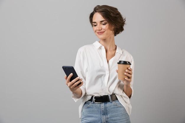 Het gelukkige jonge bedrijfsvrouw stellen geïsoleerd over grijze muur die door mobiele telefoon koffie drinken chatten