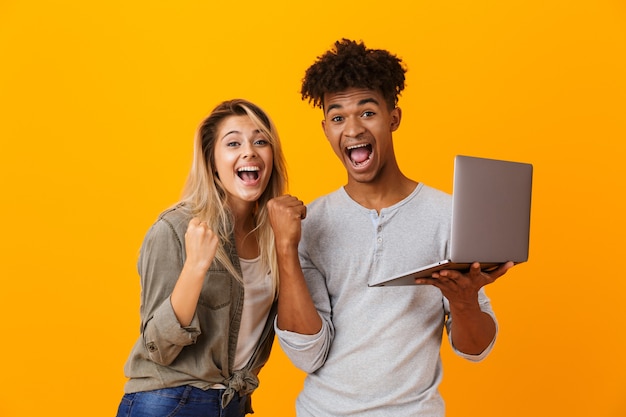 Het gelukkige jonge houdende van paar status geïsoleerd over gele muur die laptop computer met behulp van maakt winnaargebaar