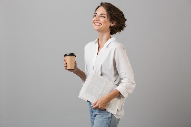 Het gelukkige jonge vrouw stellen geïsoleerd over grijze muur die koffie drinkt