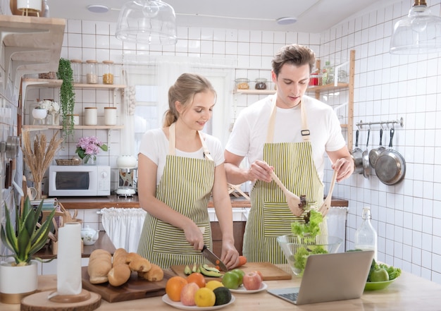 Het gelukkige Kaukasische paarfamilie koken in moderne keuken die laptop thuis met liefde met behulp van. Gehuwde romantische man en vrouw die verse groentesalade koken Gezond concept.
