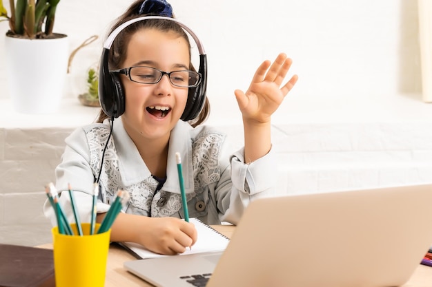 Het gelukkige meisje in een koptelefoon met een laptop die thuis aan tafel zit