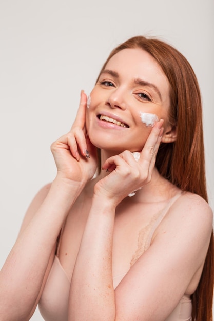 Het gelukkige mooie jonge vrouwenmodel met natuurlijk rood haar en leuke glimlach past room op gezichtshuid in studio op witte achtergrond toe Schoonheid en huidzorg