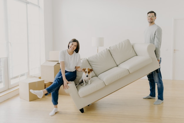 Het gelukkige paar beweegt meubilair in hun nieuw modern huis, draagt bank met huisdier, stelt in ruime ruimte