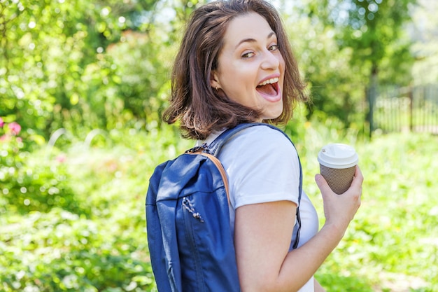 Het gelukkige positieve studentenmeisje met rugzak en haalt koffiekop het glimlachen weg