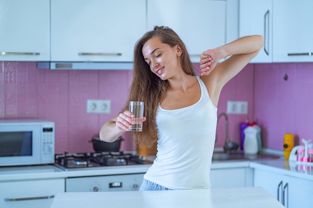 Het gelukkige slaperige vrouw uitrekken en drinkt een glas schoon gezuiverd ochtendwater in vroege ochtend na thuis ontwaken in de keuken. begin en begin van een nieuwe goede dag