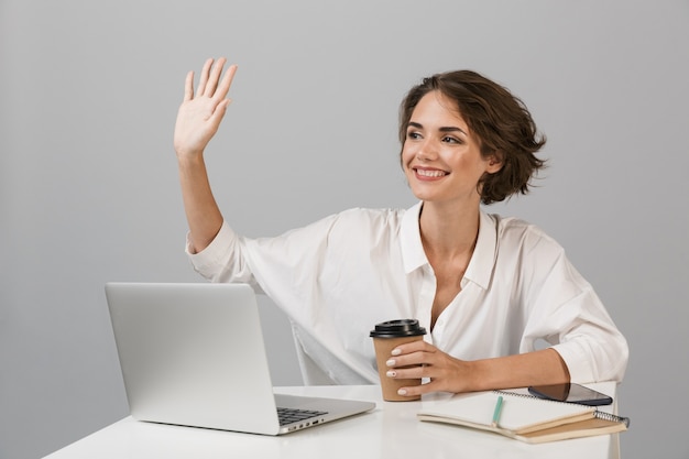 Het gelukkige vrolijke bedrijfsvrouw stellen geïsoleerd over grijze muurzitting bij de lijst die laptop met behulp van