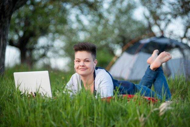 Foto het gelukkige vrouw ontspannen op het gras dat haar laptop met behulp van terwijl het kamperen in het bos