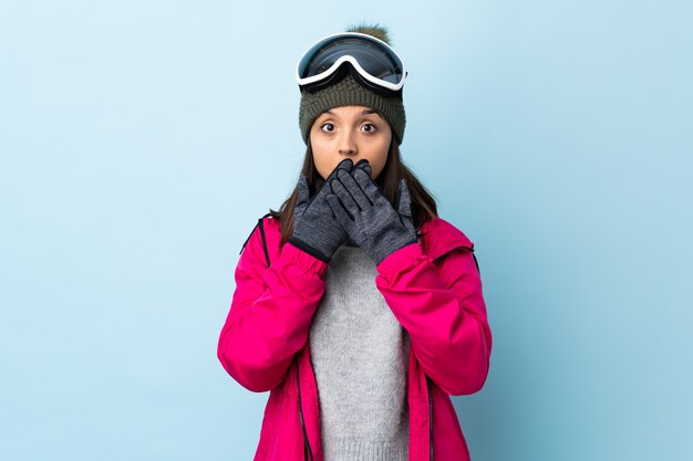 Het gemengde meisje van de rasskiër met snowboarding glazen over blauwe muur die mond behandelen met handen