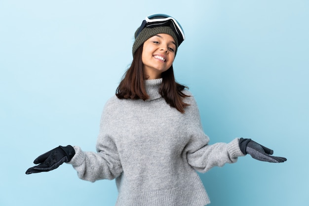 Het gemengde meisje van de rasskiër met snowboarding glazen over geïsoleerde blauwe gelukkig en muur die glimlacht