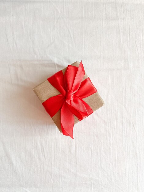 Het geschenk, vastgebonden met een rood lint en verpakt in kraftpapier, bevindt zich op een wit textieloppervlak. Kopieer de ruimte Bovenaanzicht Flatlay