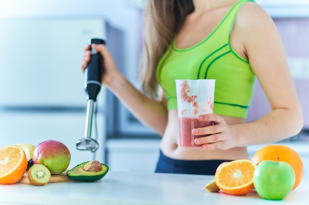 Het geschiktheidswijfje in sportkleding bereidt een vers fruit smoothie thuis met behulp van een staafmixer in de keuken voor.