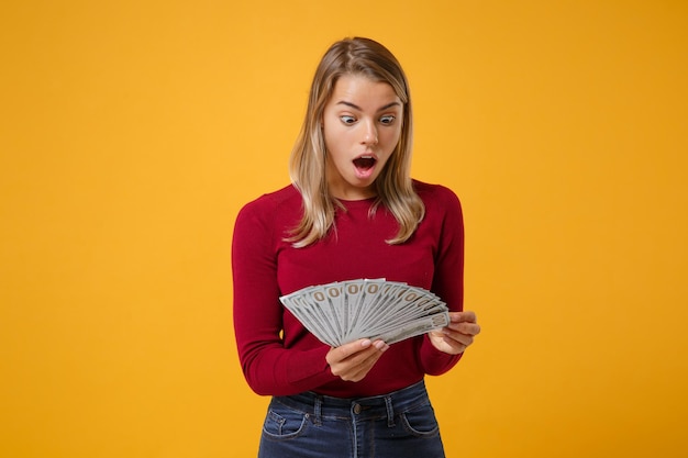 Het geschokte jonge meisje van de blondevrouw in vrijetijdskleding stellen geïsoleerd op geeloranje achtergrondstudioportret. Mensen levensstijl concept. Mock up kopie ruimte. Houd fan van contant geld in dollarbiljetten.