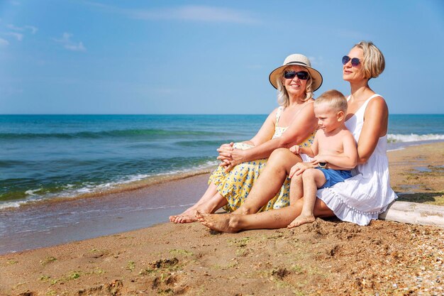 Het gezin rust op het zandstrand op een zonnige dag moeder in een witte jurk een grootmoeder in een gele zondjurk en een kleine zoon in blauwe shorts lachen en knuffelen liefde en tederheid