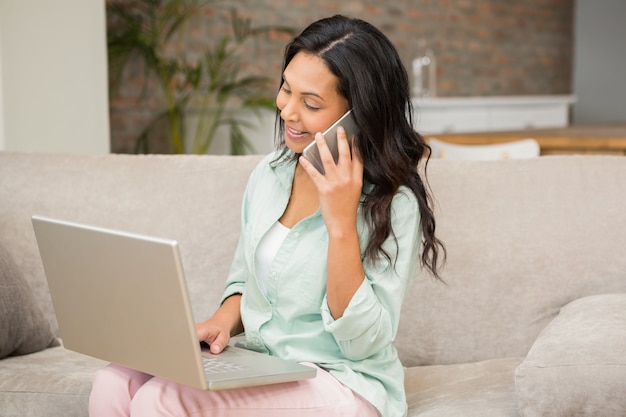 Het glimlachen brunette op een telefoongesprek die laptop op de bank met behulp van