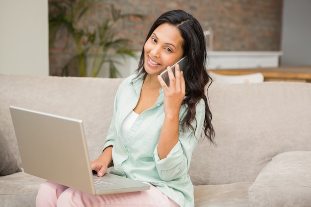 Het glimlachen brunette op een telefoongesprek die laptop op de bank met behulp van