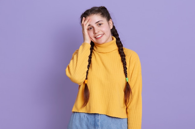 Het glimlachen van donkerharige tiener jonge vrouw bevindt zich met hand op hoofd, die gelukkige gelaatsuitdrukking hebben