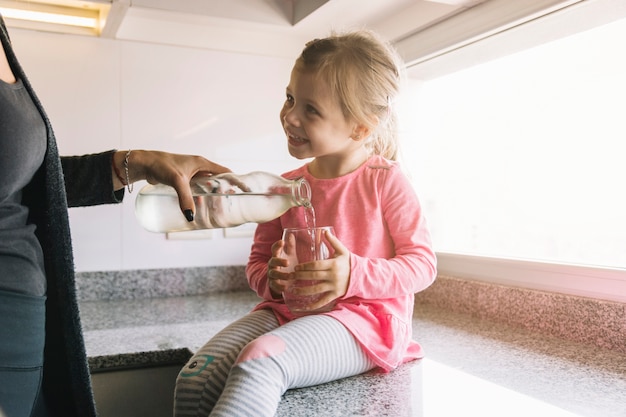 Foto het glimlachen van het glas van de meisjesholding terwijl haar moeder gietend water