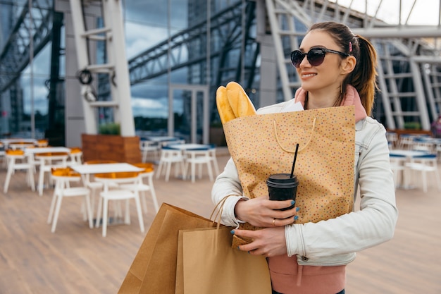 Het glimlachen van jonge de koffiekop van de vrouwenholding, die met kruidenierswinkel het winkelen document zak met lange witte broodbaguette lopen