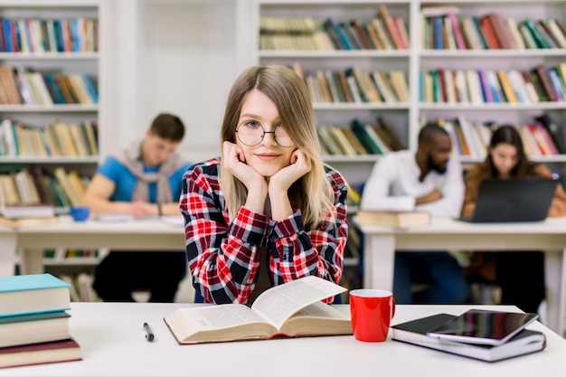 Het glimlachen van vrij jonge vrouwelijke studentenzitting bij lijst in moderne lichte bibliotheekruimte en het bekijken camera