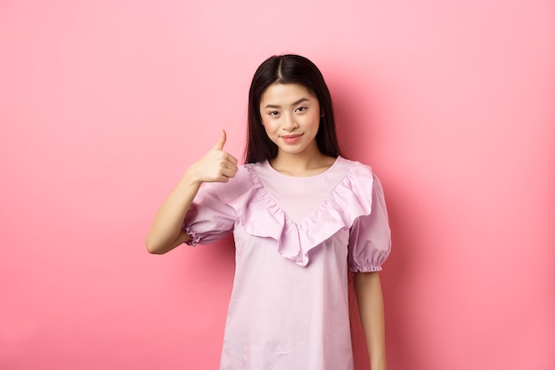 Het glimlachende Aziatische meisje toont duim ter goedkeuring, prijst de goede keuze, houdt van en beveelt het product aan, staande op een roze achtergrond.