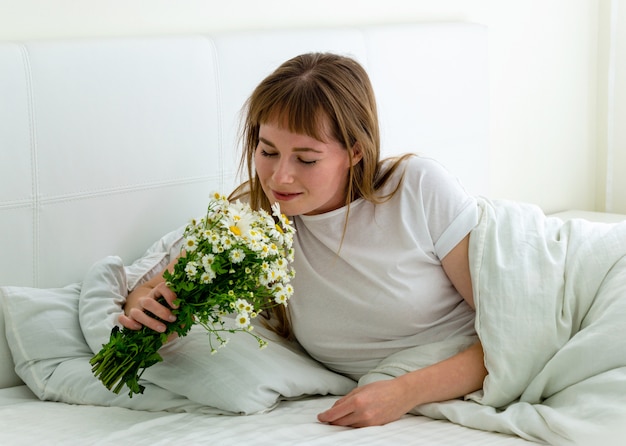 Het glimlachende jonge meisje met boeket van madeliefjes ligt thuis in bed