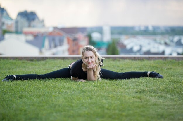 Het glimlachende jonge meisje op het groene gras voert streng op achtergrondcityscape uit