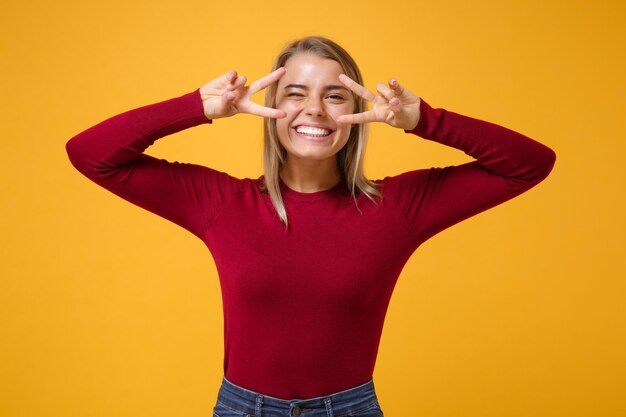 Het glimlachende jonge meisje van de blondevrouw in vrijetijdskleding stellen geïsoleerd op geeloranje achtergrondstudioportret. Mensen emoties levensstijl concept. Mock up kopie ruimte. Overwinningsteken tonen, knipperend.