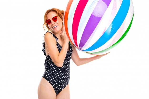 Het glimlachende meisje in retro zwempak en zonnebril uit één stuk werpt een gestreepte bal op een witte muur