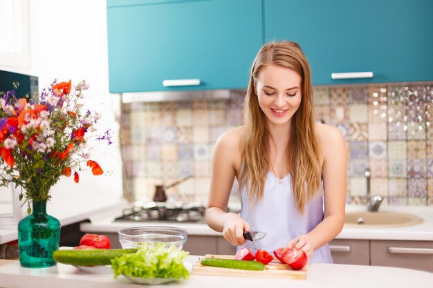 Het glimlachende mooie meisje met blond haar snijdt tomaat en komkommer voor salade op de achtergrond van heldere moderne turkooise keuken