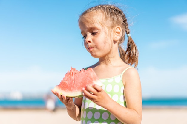 Het grappige meisje eet watermeloen zonnige de zomerdag bij oceaanstrand Leuk Kaukasisch vrouwelijk kind geniet van de beet van het zomerfruit plakje watermeloen Gelukkige jeugd