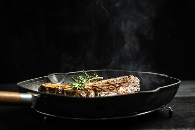 Het grillen van een lekkere malse gemarineerde t-bone steak gekruid met verse rozemarijn op een koekenpan op zwart