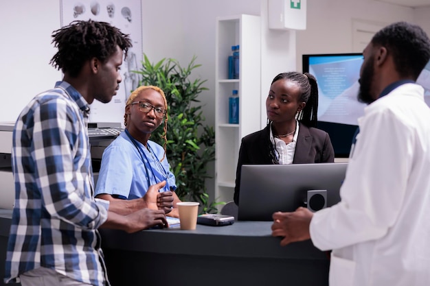 Het hele Afro-Amerikaanse team praat met de patiënt bij de receptie van het ziekenhuis om gezondheidszorg te bieden. Medisch personeel met arts, verpleegkundige en receptioniste die man met ziekte helpt in gezondheidscentrum.