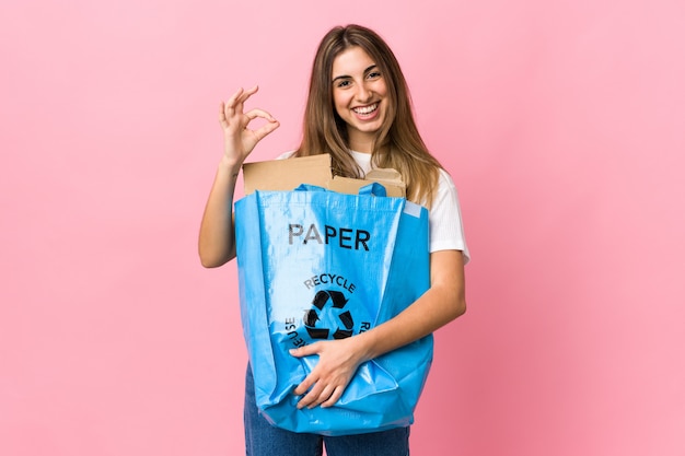 Het houden van een recycling zak vol papier te recyclen over geïsoleerde roze met een ok teken met vingers