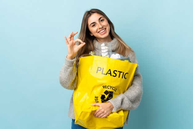 Het houden van een zak vol plastic flessen te recyclen over geïsoleerde blauw met een ok teken met vingers
