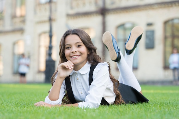 Het is weer die tijd van het jaar Ontspannen scholier Gelukkig scholier ontspannen op groen gras Klein scholier geniet van schooltijd buiten Klein scholier met lang donkerbruin haar glimlach in formele kleding