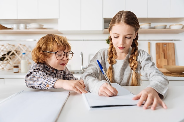 Het joch helpen. Vriendelijke, slimme, liefdevolle zus die iets opschrijft om haar broertje te helpen met zijn thuistoewijzing terwijl ze allebei thuis zijn