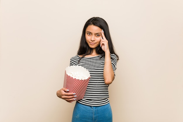 Het jonge Aziatische meisje die een popcornemmer houden richtend zijn tempel met vinger, denken, concentreerde zich op een taak.