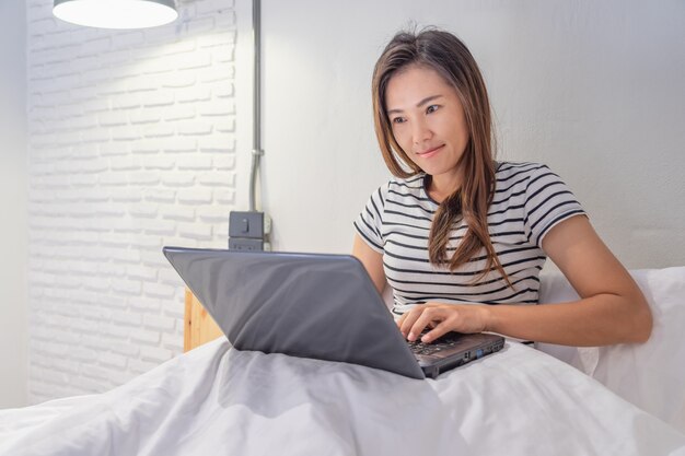 Het jonge Aziatische vrouw spelen met laptop op haar bed in witte slaapkamer